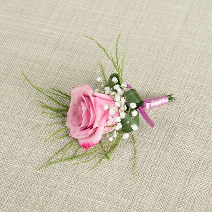A delicately crafted pink rose boutonniere, suitable as a traditional buttonhole or a petite corsage, lies on a neutral beige linen backdrop, the fabric's fine weave adding a quiet, organic texture reminiscent of the table linens in small Canonbury venues and cafés. Viewed from directly above, the arrangement is compact yet detailed, centred on a single rose whose petals are numerous and tightly layered in the middle, then gradually loosen and open as they spiral outwards. The colour progression is gentle but distinct: a warm, soft pink at the heart that slowly deepens into a more intense magenta along the outermost petal edges, each petal slightly ruffled to suggest a fresh, recently opened bloom. Nestled closely around the rose are clusters of white baby's breath, each spray composed of tiny, star-like blooms that together form a misty, cloud-like ring. These miniature flowers provide a crisp, white contrast that brightens the overall composition and underscores the rose's colour. Two complementary types of greenery frame the flowers: fine, bright green grass-like fronds extend from beneath the rose in graceful, radiating lines, adding a sense of movement and airiness, while a single broader, darker leaf has been carefully folded and wrapped around the base, creating a structured, almost cuff-like cradle for the stems. The lower portion of the boutonniere is neatly finished with a slim, light pink satin ribbon, wrapped smoothly around the stems and tied into a small, symmetrical bow with short, tidy tails that lie flat against the linen. Subtle, even lighting ensures that no part of the arrangement is harshly shadowed; instead, the curves of the petals, the rounded shapes of the baby's breath, and the polished bow are all softly defined. This refined piece feels perfectly at home pinned to a lapel for a Canonbury wedding, civil ceremony in nearby Islington, or an elegant prom night, offering a quietly romantic accent that photographs beautifully against suits and dresses alike.
