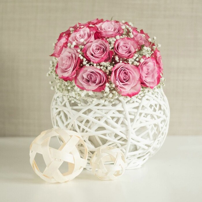 An artfully styled tabletop scene features a lush, spherical bouquet of pink roses and delicate white baby's breath, the kind of arrangement a local Canonbury florist might set in the window of a shop off St Paul's Road. The bouquet is densely packed, forming a full, rounded silhouette, with each rose carefully angled so its open face contributes to the overall dome. The petals exhibit a graceful gradient, starting with a soft, powdery pink at the centre and gradually intensifying into richer, almost fuchsia tones along the outer edges, giving each flower a sense of depth and maturity. Between the roses, fine stems of Gypsophila thread through, their tiny white blossoms appearing like a dusting of snowflakes or a veil of light mist. This baby's breath softens the transitions between blooms and creates pockets of airy texture that make the composition feel lighter and more ethereal. The bouquet rests inside a distinctive spherical vase, crafted from white-painted rattan or a similar natural fibre, woven into an open basket-like lattice. The structure allows glimpses of shadow within, while the exterior catches the soft, even light that falls across the scene, creating gentle highlights on the raised strands. On the gleaming white surface in front of the vase, two decorative spheres made from slender, pale wooden strips echo the weave of the container. One is noticeably larger than the other, and their placement to the left adds balance and a sense of curated minimalism. Behind everything, a warm beige or off-white wall, subtly textured, forms a calm, neutral backdrop. The atmosphere is peaceful and stylish, ideal for illustrating a romantic gift, a Mother's Day surprise, or a refined home decor idea for residents across Canonbury and nearby Islington.