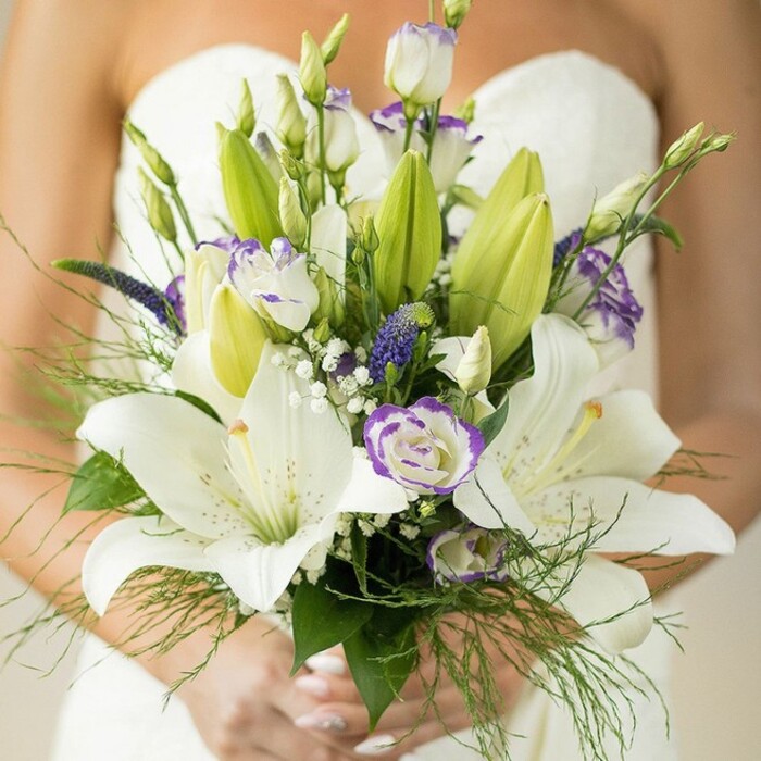 In the heart of Canonbury, this breathtaking bridal bouquet is carefully held by a bride in a classic strapless white gown, exemplifying timeless London elegance. The photography captures every subtle detail: large, snow-white Oriental lilies, each flower unfurling with delicate spotted petals and bold yellow-orange stamens, some still wrapped in pale green buds. Between the lilies, clusters of Eustoma (lisianthus) bloom-some petals gently crinkled, some edged in a painterly wash of soft lavender-purple, exuding gentle vibrancy and softness. Slender spikes of deep amethyst or indigo-purple, suggestive of Muscari or Veronica, add dramatic accents echoing the colors of nearby Regent's Canal wildflowers. Sprays of traditional white baby's breath (gypsophila) form airy layers, while lush greenery envelops the bouquet-broad, deep green leaves and airy, fern-like foliage cascading with effortless grace. The bride's hands, their nails painted a pale neutral, delicately cradle the arrangement-her dress and bare shoulder softly blurred behind, focusing all attention on the florals themselves. The bouquet's sensuous blend of colour, movement, and texture radiates romance, purity, and luxurious local craftsmanship. Expertly curated for a wedding in Canonbury or across Islington, it is a signature creation worthy of magazine spreads and community celebrations alike.