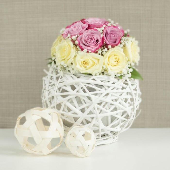 The image presents an elegant floral centerpiece that would feel perfectly at home on a wedding table in Canonbury or a stylish dining room just off Essex Road. At the centre is a tightly gathered, almost spherical bouquet of roses, their heads crowding together to form a plush, domed cushion of petals. Around a dozen blooms are visible, half of them in a delicate pastel pink with hints of lavender, the others in a warm ivory and soft butter-yellow, creating a soothing, romantic contrast. The roses are in different stages of opening: some remain gently cupped, others are already unfurling, revealing intricate layers that catch the light. Interlaced among the roses are slender stems of white baby's breath, each tipped with clusters of tiny blossoms that soften the edges of the bouquet and add an airy, cloudlike texture. This floral crown rests in a distinctive white spherical vase, created from interwoven twigs or wicker painted in a crisp white. The strands cross and loop around one another, forming an open lattice that gives a glimpse of the container hidden within, and lends the piece a rustic-modern charm that would suit many Canonbury terraces. The vase sits on a smooth, pure white table surface, its simplicity drawing attention to the craftsmanship of the arrangement. To the left, two decorative spheres echo the wicker texture, though they are made from natural, light-toned wood or rattan, one noticeably larger than the other, balancing the composition. The background is a soft, neutral beige, with a subtle woven or fabric-like texture that suggests a refined, cosy interior. Bathed in gentle, even light, the scene feels calm, welcoming, and thoughtfully styled, ideal for inspiring wedding décor, anniversary dinners, or sophisticated home styling in the Canonbury area.