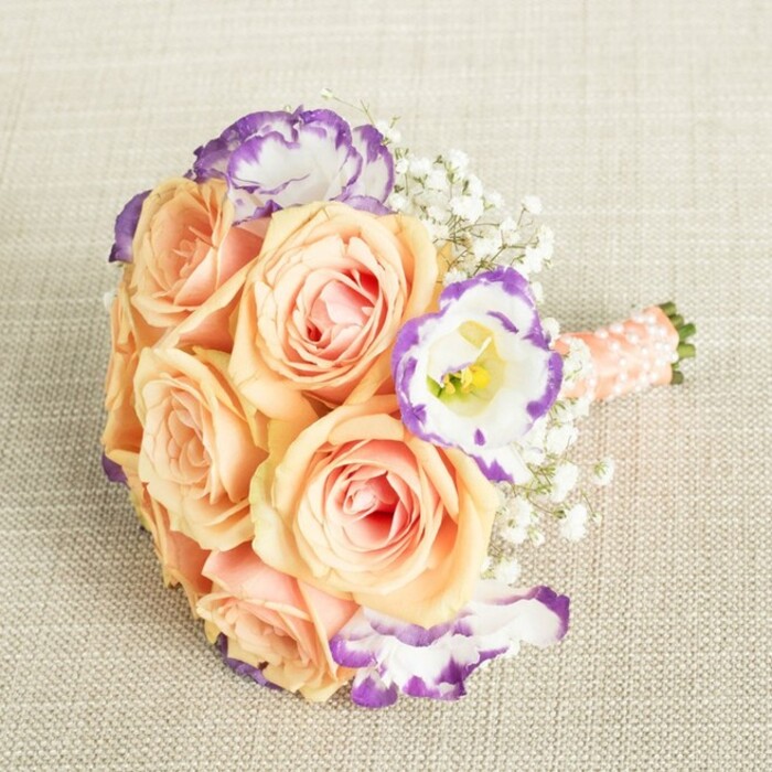 Lying serenely on a textured beige cloth, a bouquet reminiscent of Canonbury's elegant charm catches the eye and imagination. Roses in soft peach, apricot, and cream form the lush core, their petals opening to blush centers as if unfurling under the natural light from a studio window overlooking the Regent's Canal. Sprinkled among them, Lisianthus blooms unfold, their ruffled white petals edged in vivid purple, painting subtle drama into the soft pastel composition. Airy Baby's Breath floats between the roses, softening each edge with cloud-like fullness. Every stem is carefully wrapped in creamy peach-pink satin, adorned neatly with a trail of pearl beads-details that echo craftsmanship typical of Canonbury Flowers, with hints of romance found in Barnsbury's hidden gardens. The bouquet's balanced, rounded shape cradles each element-ideal for clutching on a wedding day, gracing a family celebration, or bringing a moment of refined beauty into a home near Upper Street. This arrangement tells a gentle local story, its every nuance conveying thoughtful artistry, joyful anticipation, and the freshness only a beloved Canonbury florist can provide.
