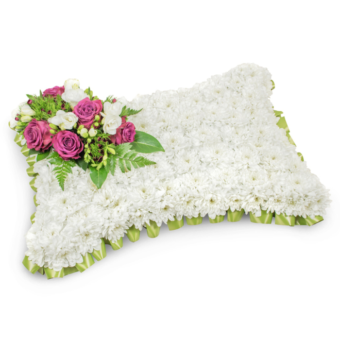 A carefully formed pillow of flowers rests against a pristine white field, its rectangular form defined by a continuous carpet of small, immaculate white chrysanthemums that produce a uniformly plush texture reminiscent of a soft linen cushion. The arrangement's upper left corner carries an accent cluster composed of several rich magenta and fuchsia roses; their layered petals open with a velvet quality that provides a poignant contrast to the surrounding white. Interspersed among these roses are sprays of diminutive white freesias or comparable buds, offering both fragrance and a delicate, lace-like counterpoint. Supporting foliage - narrow fern fronds and assorted glossy greens - fans beneath the focal grouping, lending depth, natural contour and a cooling verdant backdrop. The outer edge of the pillow is neatly finished with a pleated or looped trim of pale green satin ribbon that peeks out beneath the chrysanthemums, giving the rectangular silhouette a finished, ceremonial touch. Subtle studio lighting reveals the tactile gradations across the surface and a soft shadow below that accentuates three-dimensional form without distracting from the composition's serene intent. Designed for memorials and funeral services, this traditional pillow arrangement embodies repose, respect and tender remembrance; it is the kind of bouquet families in Canonbury and neighbouring streets might choose to symbolise eternal rest, offering a composed, comforting visual tribute during times of bereavement.