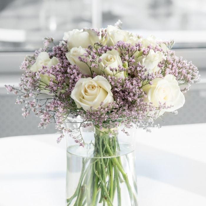 In this softly lit photograph, a refined bouquet of creamy white roses and delicate purple statice is captured in intimate detail, expressing the kind of calm reassurance often sought in times of loss. A cluster of open white roses commands the eye, each blossom formed from layers of velvety petals that curl gently back from a tight spiral centre. Their tones shift subtly from pure white at the outer edges to a faint ivory glow near the heart of each flower, creating a sense of depth and quiet luxury without feeling showy. Interwoven among the roses are abundant sprigs of tiny lavender and pale lilac filler flowers, likely statice, whose many small, papery blooms create a soft, clouded halo around the larger heads. A hint of blush pink occasionally touches the filler, adding just enough nuance to keep the palette from feeling stark. The stems, neatly gathered and trimmed, stand straight within a tall, cylindrical glass vase filled with fresh, clear water, every line visible through the glass like a simple, honest framework holding the arrangement together. The vase is placed on a bright white surface that reflects the gentle daylight, reminiscent of the natural brightness you might find near Canonbury Square or in a quiet home off St Paul's Road. Behind the bouquet, the background fades into a blur of very pale greys and whites, reinforcing the sense that this bouquet is the single, thoughtful focus of the moment. The overall impression is of a carefully crafted condolence arrangement, suitable for a remembrance gathering, a funeral service, or a private gesture of sympathy delivered to a family home anywhere across Canonbury and surrounding North London neighbourhoods.