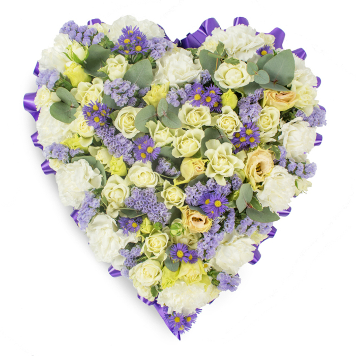 An elegant heart-shaped sympathy arrangement captured on a plain white background, where delicate creams and soft lavenders mingle to form a soothing, heartfelt composition. At the core are numerous cream and pale yellow roses - some in tight bud, others gently opening - their petals layered to give a plush, sumptuous texture that reads as tender and dignified. Interspersed throughout are full, ruffled white carnations which add classic body and a comforting familiarity, while sprays of lavender-hued statice lend a fine, paper-like texture and a whisper of wildflower charm. Pockets of small purple, daisy-like asters with vivid yellow centres create bright, optimistic moments against the softer tones, providing visual punctuation and a touch of liveliness. Rounded, blue-green eucalyptus leaves are scattered with intentionality, their cool, muted colour and smooth edges calming the composition and suggesting the faint, green scent of crushed leaves. A continuous, rich purple satin ribbon loops around the heart's edge, neatly folded to produce a refined frame that defines the silhouette and elevates the tribute. The soft studio illumination brings out subtle shadows and highlights across petals and foliage, enhancing the sense of depth and care in the arrangement. As Canonbury Flowers, crafting tributes for services near Canonbury Square or St Paul's, we choose this palette to convey peace, respect, and enduring affection - a dignified memorial piece meant to comfort mourners and honour a beloved life with quiet, heartfelt beauty.