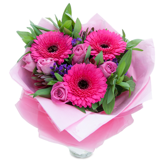There is a playful grace to this hand-tied bouquet that feels both intimate and refined. Three prominent fuchsia Gerbera daisies open like cheerful faces, their saturated petals creating movement and a confident centre of attention, while nearby roses in a softer lavender-pink or dusty rose provide a gentle, romantic counterpoint with their velvety, inward-turning petals. Fine sprays of purple statice add a whimsical, meadow-like texture between the larger blooms, their tiny clustered blooms offering a cool blue-purple whisper against the warmer pink tones. The foliage beneath and around the flowers is full-bodied and verdant-broad leaves that read as salal or eucalyptus, slightly glossy and tactile, bringing a crisp, green freshness that both supports and showcases the petals. The bouquet is wrapped with care: an inner wrap of pale pink tissue cradles the stems and softens the presentation, while an outer layer of sheer, crinkled fuchsia-pink cellophane adds a soft sheen and helps the colours sing together; the top rim of a clear glass vase is visible at the base, suggesting immediacy and readiness to display. Under natural daylight the colours glow warmly, and the mixed textures-from papery statice to plush rose petals-create a pleasing contrast to touch and sight. Perfect for placing on a bedside table or sending as a cheering gift to Newington Green or Chapel Market neighbours, this bouquet radiates affection and thoughtful craftsmanship, the sort of arrangement a local florist would make to celebrate small, meaningful moments.