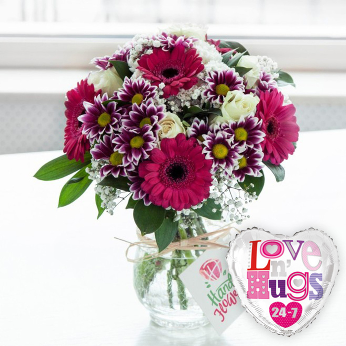In this bright, uplifting scene, a joyful bouquet and a heart-shaped balloon come together to create the impression of a surprise delivery arriving at a Canonbury doorstep. The flowers are arranged in a generously rounded style, led by several large gerbera daisies in rich fuchsia and magenta tones, their broad petals arranged in perfect concentric rings around distinctive dark centres. Nestled closely around them are clusters of striking chrysanthemums whose purple petals fade delicately to white at the edges, each bloom centred with a vibrant yellow eye that glows against the deeper hues. Cream-toned white roses are tucked thoughtfully throughout the design, some still in tighter buds, others just beginning to open, giving a sense of gentle movement as if the bouquet is unfolding. Wisps of fine white baby's breath thread between the larger blossoms, creating a light, gauzy veil that softens the strong colours. Fresh green leaves and foliage emerge from the base and around the sides, adding both structure and a natural, garden-fresh character typical of designs crafted by Canonbury florists for local homes and studios. The arrangement is presented in a classic clear glass vase that reveals the neatly aligned green stems and clear water inside, suggesting care and freshness. Around the narrow neck of the vase, a piece of rough-textured natural twine is tied and finished with a small white tag, illustrated with a pink rose icon and handwritten-style green lettering referencing the handmade nature of the flowers. Just to the right, touching the edge of the vase, a shiny silver-bordered foil balloon in the shape of a heart adds a playful note, its white centre filled with colourful, patterned letters spelling a warm Love n' Hugs 24-7. Resting on a light, uncluttered surface with a softly blurred, sunlit background that could be a window looking toward Islington's quiet streets, the image conveys a sense of celebration, affection, and the delight of receiving flowers at home or work.