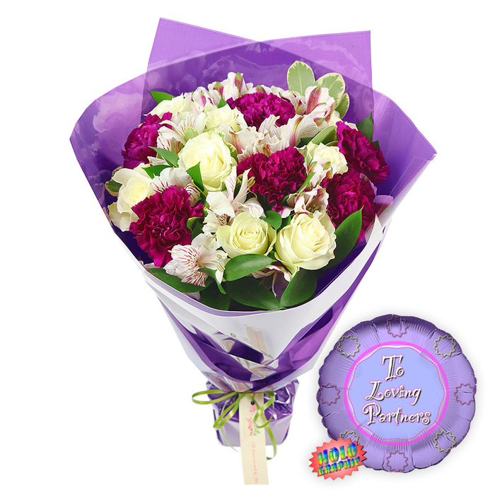 A tender, celebration-ready bouquet with a coordinating balloon rests against a pure white background, the kind of display you might see carefully prepared for collection in Canonbury on a special anniversary afternoon. The bouquet itself is rich and full, dominated by creamy white roses whose tightly layered petals form elegant spirals, each bloom suggesting purity and devotion. Interlaced with them are numerous deep magenta and burgundy carnations, their compact, frilled petals creating lush, cushiony textures that radiate warmth. Adding a lighter, more intricate note, white alstroemeria-Peruvian lilies-arc through the arrangement, their slim petals painted inside with fine reddish-brown stripes and speckles that look almost hand-drawn. Surrounding and supporting these focal blooms, fresh greenery fans out in varied shapes and tones: glossy deep green leaves, softer foliage with pale, variegated margins, and small sprigs that break up the colour fields, all helping the purples and creams stand out. The florist has wrapped the stems in a sophisticated two-layer combination: closest to the flowers lies a smooth, angled band of white paper, offering clean lines and gentle structure, while the outer layer is a translucent purple cellophane or film. That purple wrapper rises behind and above the heads of the blooms, slightly crinkled, catching reflections and giving a soft, glowing aura around the bouquet. At the gathered base, a slim bright green satin ribbon is tied into a petite bow, with a small light brown card tag slipping from the knot, suggesting a handwritten note from someone in Canonbury or nearby De Beauvoir. Beside the bouquet, to the right, sits a small round metallic purple balloon, its edge scalloped like a decorative rosette. In the paler lilac centre panel, the words To Loving Partners appear in graceful script, while a tiny rainbow holographic sticker clings to the lower side, hinting at a shimmering finish. The ensemble conveys romance, tenderness, and shared celebration-perfect for marking relationship milestones or quiet evenings together in Canonbury homes.