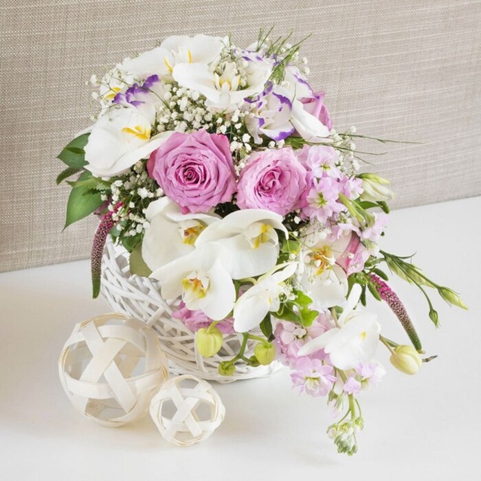 This image presents a beautifully detailed floral centerpiece, photographed at close range so each petal and woven fibre can be appreciated, as though the flowers were set on a wedding table in a quiet Canonbury townhouse. A round white wicker basket forms the base, its tightly interlaced strands creating a rustic yet polished container that contrasts gently with the soft, organic shapes of the flowers. Rising from the basket is a generous arrangement in shades of blush pink, crisp white, and muted lavender. Plump pink roses sit prominently at the centre, their perfectly spiralled petals wide open, suggesting both abundance and romance. Interspersed with the roses are striking white Phalaenopsis orchids, their broad, satin-like petals and golden-yellow centres giving an immediate sense of luxury, as if chosen for a sophisticated celebration at a nearby Islington venue. Around these focal blooms, white lisianthus blossoms, some edged in a subtle purple hue, add layers of texture and a slightly ruffled softness that keeps the design from feeling too formal. Long stems of pale pink stock gently overflow the basket's rim, providing height and a graceful, cascading line that draws the eye outward. Sprays of delicate white baby's breath are tucked among the larger flowers, lightening the overall look and creating tiny points of interest that resemble stars scattered through the arrangement. Fresh green foliage peeks out all around, giving the impression that the flowers have just been gathered and arranged by hand. On the smooth white surface in front of the basket, two decorative woven orbs in natural wood tones echo the basket's material and add sculptural interest. A light beige, linen-textured backdrop softly frames the arrangement, allowing the gentle colours to shine while conveying a warm, intimate mood suitable for describing local Canonbury wedding flowers or special event centrepieces.