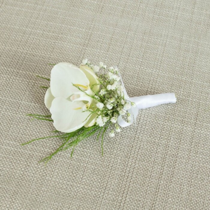 A graceful white orchid boutonniere is captured in calm, natural light as it rests on a light beige backdrop, the woven fabric surface recalling the understated, contemporary textiles often seen in Canonbury's elegant homes. At the centre of the composition, a single Phalaenopsis orchid bloom unfurls its broad, curved petals in an almost sculptural form, the white appearing clean and cool with just a whisper of cream and a faint glow of pale yellow at the throat. This inner detail draws the eye inward and hints at the bloom's freshness, as though it has only recently been wired and placed by a skilled local florist. Framing the orchid is a ring of baby's breath, delicate Gypsophila blossoms clustered on hair-fine stems, creating a soft, cloud-like aura around the central flower. Each tiny, white bloom adds texture and volume without overwhelming the orchid, lending the boutonniere a romantic, weightless quality reminiscent of a gentle mist drifting across nearby New River Walk. Slender, feathery sprigs of bright green foliage, likely a fine fern or similar greenery, emerge from beneath and around the orchid, their narrow fronds adding a sense of organic movement and a refreshing colour contrast. The stems are gathered neatly and wrapped in a smooth white satin ribbon, the wrap clean and symmetrical, finished with a small, precise bow that rests just above the cut ends. This careful binding suggests professional craftsmanship, the kind associated with dedicated shops like Canonbury Flowers, which often prepare such pieces for intimate townhouse ceremonies and chic London receptions. The overall mood is one of quiet sophistication and modern simplicity, a timeless floral accent suited for a groom, best man, or guest attending a wedding or formal celebration across Canonbury and the surrounding Islington neighbourhoods.