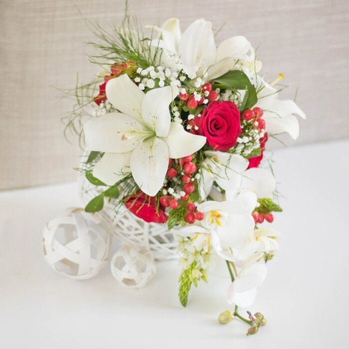 An elegant wedding-inspired floral arrangement is showcased on a crisp white tabletop, set in front of a gently textured beige backdrop that recalls the calm, muted interiors of Canonbury terraces. The flowers are cradled in a round white basket-style container, crafted with a delicate open lattice that lets light move through it, giving the whole display a weightless, artisanal character. Rising from this airy base, several large white oriental lilies command attention, their petals smooth and luminous, curving outward to reveal tall stamens dusted with a hint of pollen. Intertwined between the lilies, velvety red roses in a deep, romantic shade unfold in varying stages of bloom, from tightly wound buds to fuller, cup-shaped flowers, bringing richness and passion to the palette. Fine sprays of white gypsophila, or baby's breath, drift softly throughout, creating little pockets of haze that soften the bold lines of the main blooms. Bright green, feathery foliage reminiscent of fern adds texture and depth, extending gently beyond the basket's rim, as though freshly gathered by an experienced Canonbury florist for an intimate reception along Essex Road. Dotted between the flowers, clusters of glossy red hypericum berries offer small, jewel-like accents, their rounded forms echoing the smooth curves of the roses. From the right side, a graceful strand of white Phalaenopsis orchids cascades outward, its slender stem carrying multiple elegant blooms with delicate yellow and blush pink centres, along with a few tightly closed buds at the tip, suggesting ongoing life and continuity. On the left of the main arrangement, three white decorative spheres made from the same woven material rest in a loose triangle on the table, mirroring the basket's texture and visually anchoring this sophisticated, romantic floral composition.