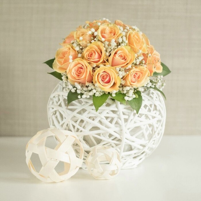 A lush, dome-shaped bouquet of peach and apricot roses is captured here as the focal point of an elegant indoor scene, the kind you might find styled on a console table in a Canonbury townhouse. Around twenty to thirty rose heads cluster tightly together, creating a rounded crown of colour. Each flower shows off layers of satin-like petals: the centers glow with deeper orange and warm apricot tones, while the outer petals fade into softer, powdery peach, lending the arrangement a gentle, almost sunrise-like gradient. Nestled among the roses are airy bunches of white baby's breath, their tiny, cloud-like blooms scattering light and adding a sense of movement. Tucked at the base, small glimpses of dark green foliage add depth and a natural frame, anchoring all the softness. The bouquet sits in a distinctive, spherical vase made of white woven strips, reminiscent of wicker or rattan, arranged in a loose lattice that feels both modern and organic. The open pattern allows light to pass through and cast subtle shadows on the bright white tabletop below. In front of the main vase lie two decorative orbs, crafted from pale, natural-looking strips woven into open, geometric spheres. One is slightly larger, one smaller, and together they echo the textural play of the vase without competing with the flowers. The background is a softly textured light beige wall, its vertical lines faint but present, providing a calm, neutral stage that lets the warm roses and crisp whites stand out. Bathed in even, natural daylight, the scene suggests quiet luxury and thoughtful design, perfectly suited to a wedding reception at a local Canonbury venue or an intimate celebration in a refined Islington home, where fresh flowers are used as a subtle expression of warmth and welcome.