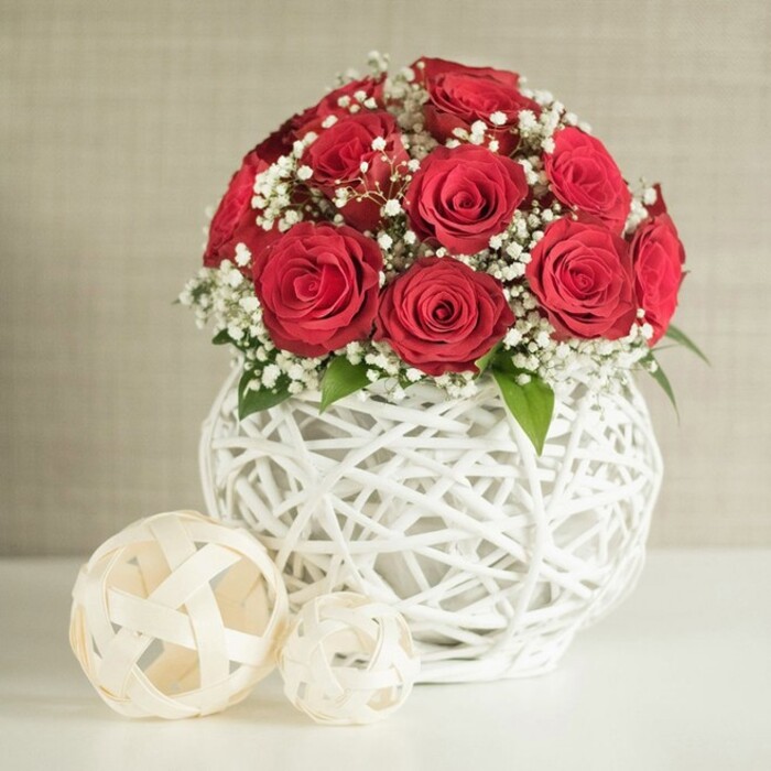 A rich, spherical cluster of deep red roses sits proudly in the centre of the frame, designed with the kind of precision you might expect from a wedding table set for guests in Canonbury's elegant townhouses. There are roughly a dozen to fifteen roses, each one open at its prime, with tightly layered petals forming lush, velvety cups of colour. No bloom crowds another; instead they nestle closely, creating a seamless dome of crimson that feels both opulent and calm. Interwoven among the roses are generous sprigs of baby's breath, their tiny white stars sprinkled like light snowfall across the arrangement. These delicate blossoms soften the strong red, while a few discreet green leaves emerge from beneath, lending depth and a whisper of garden freshness, as though the bouquet had been gathered just moments ago from a hidden courtyard near Canonbury Place. The flowers are cradled in a round white vase with a distinctive open lattice pattern, crafted from intertwined strands that resemble painted wicker or rattan. The structure allows glimpses of light and shadow to play through the gaps, adding an airy, handcrafted feel. On the smooth white tabletop, slightly to the left of the vase, rest two decorative woven spheres in an off-white, natural tone. The larger ball leans subtly toward the viewer, with the smaller one nestled behind, creating a gentle sense of perspective. A softly textured, neutral wall forms the background, blurred enough to keep all focus on the arrangement. The overall atmosphere is one of serene sophistication, ideal for a wedding reception, anniversary dinner, or intimate celebration in a quiet Canonbury dining room.