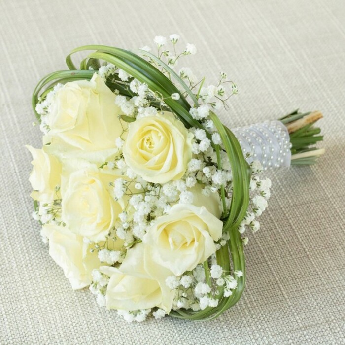 Expertly styled atop a softly textured beige linen reminiscent of classic Canonbury interiors, this bridal bouquet offers a vision of refined beauty. Approximately eight roses take the spotlight-their creamy white and pale yellow petals artfully unfurling, each bloom reflecting the tranquility of the nearby New River Walk. Nestled all around are voluminous clusters of delicate, white gypsophila, their cloud-like forms providing an ethereal foil to the substantial roses. Intertwined, loops of slender green bear grass impart lift and modern edge, reminiscent of the decorative greenery spotted in local Islington florists' window displays. Every stem is gathered in a sleek, silver-grey satin ribbon, painstakingly accented with three rows of lustrous white pearls, delivering a sense of luxury and tactile charm fit for any special celebration. The fresh green stems, neatly trimmed, peek just beyond the ribbon's edge. This bouquet's artistry and nuanced textures embody the classic romance and timeless sophistication cherished in Canonbury's vibrant wedding scene-perfect for elevating ceremonies, anniversaries, or cherished gatherings across north London.