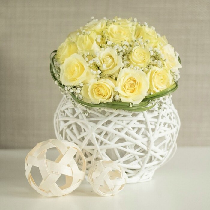 An exquisite wedding table arrangement is showcased against a soft, neutral backdrop, its gentle glow reminiscent of early evening light drifting through a Canonbury terrace window. At the centre of the scene, a rounded bouquet of creamy yellow roses rises like a small sun, each flower fully open and beautifully formed. The petals, velvety and full, overlap in tight spirals, displaying subtle variations of buttery yellow and pale cream that add depth and movement. Between the roses, fine sprigs of baby's breath create a delicate veil, the Gypsophila's clustered white blossoms lending a cloud-soft texture that feels almost weightless. Just at the base of the domed bouquet, a slim band of fresh green leaves is wrapped with precision, as if gently cinching the flowers together and lending a tailored, polished finish to the design.   

The arrangement stands in a distinctive white woven vase, whose structure is reminiscent of rattan or wicker yet rendered with a modern, sculptural twist. Its open latticework curves gracefully, wider at the top and lower edge, then drawing in slightly at the centre like a contemporary urn. Light filters through the gaps, creating slender shadows on the glossy white tabletop and adding subtle visual rhythm. Beside the vase sit two decorative woven spheres, crafted from pale, natural-looking strips of wood or similar material. Their open, crisscross structure mirrors the vase's texture, uniting the elements into a calm, cohesive composition. The larger ball is set toward the foreground, the smaller one nestled nearby, as though thoughtfully positioned for a reception setting in Canonbury, perhaps overlooking paths that lead down toward Essex Road or the Regent's Canal. The softly blurred beige background ensures no distractions, allowing the viewer to focus on the gentle interplay of yellow, white, and green. The overall mood is one of refined joy and intimate celebration, ideal for wedding breakfasts, anniversary dinners, or elegant gatherings in Islington's characterful venues.