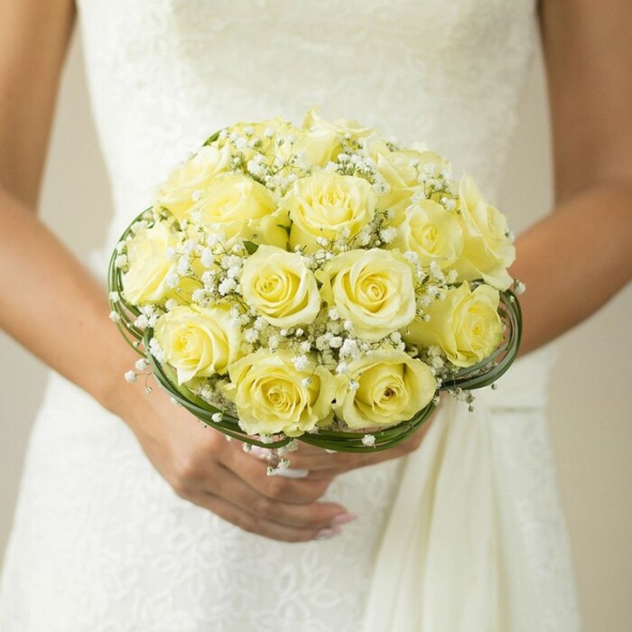 In a beautifully composed close-up rooted in the charm of Canonbury, a bride, seen from the shoulders down, cradles a hand-tied bouquet designed with thoughtful care. The bouquet's heart is a tight cluster of soft yellow roses, their petals elegantly curled and glistening faintly, forming a lush circular crown. Each rose, perfectly open and fresh, contrasts gently against the subtle ivory patterns and textures of her white gown, possibly bearing lace reminiscent of Canonbury's period homes. Airy, snow-white sprays of baby's breath are intricately interspersed, lending a delicate movement and traditional accent to the bouquet. Fine, dark green leaves or whisper-thin bands of decorative wire trace around the base, introducing a stylish frame that accentuates the richness of the blooms. The bride's manicured hands, topped with a newly placed wedding band, support the flowers with reverence, epitomizing the joy and anticipation of matrimony. Soft focus and diffused light highlight the central arrangement, much like the ambience in a local wedding at New River Walk or Canonbury Square. This evocative image embodies celebration, romance, and the artistry found within every floral creation made for North London brides.