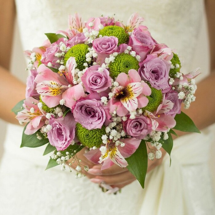 Positioned intimately in the bride's gentle hands against the refined texture of her lace-trimmed gown, this vibrant Canonbury bridal bouquet captures the eye and the heart. At the centre, blossoming roses shift from soft lavender to delicate pink, a natural unfolding that whispers romance and delicacy. Dew-kissed petals glisten subtly in the light, recalling fresh market mornings on Canonbury Road. The vibrant, pom-pom-like green button chrysanthemums add a breezy, uplifting contrast, infusing the bouquet with a sense of playful modernity. Tucked amidst these are pink alstroemeria lilies, their petals marked with distinctive maroon stripes and hints of golden yellow, imparting an exotic yet refined energy. Filigrees of white baby's breath cocoon the arrangement, contributing a lace-like airiness and a cloud-soft finish, reminiscent of early spring blooms seen in local gardens. Finishing the bouquet, rich green leaves embrace the overall design, reinforcing its sculptural fullness. Every detail speaks to the artistry of Canonbury florists-perfect for North London couples envisioning a wedding imbued with romance, freshness, and elegant, contemporary style.