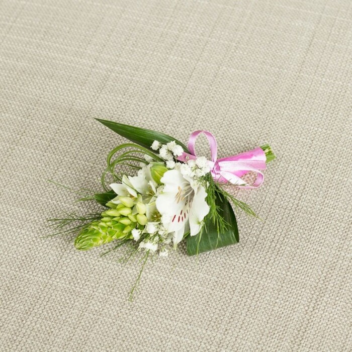 A small yet artfully composed floral piece is displayed on a subtly textured, light beige cloth that resembles finely woven linen, the neutral backdrop allowing each detail to stand out with clarity. At the centre sits a single white alstroemeria, also known as Peruvian lily, its petals gently flaring outward in a soft star. The petals are smooth and slightly ruffled at the tips, with distinct reddish-brown speckles gathered near the throat, lending the flower both intricacy and character. Just to one side rises a vivid lime-green spire of tightly packed buds, an elongated cone that feels almost sculpted, providing a bold textural counterpoint to the alstroemeria's open softness. Scattered among these main elements are fine sprays of baby's breath, their tiny, cloud-like white blossoms perched on thin stems, filling the gaps with a delicate haze that softens edges and adds a romantic veil to the design. Supporting the flowers is a foundation of lush foliage: broad, dark green, pointed leaves fan outward like small blades, while slender strands of bright green, grass-like foliage loop and curve, suggesting movement and giving the arrangement a natural, garden-picked charm. The stems are neatly bound with a narrow band of glossy pink satin ribbon, tied into a precise, compact bow that introduces a lively note of colour and a hint of playful elegance. The lighting in the image is even and gentle, with no harsh contrasts, so every petal vein and leaf edge can be appreciated. This refined boutonniere or small corsage feels ideal for a Canonbury wedding party heading to a ceremony near Islington Green or a formal celebration in one of the area's Georgian venues, offering a subtle accent that speaks of care, craftsmanship, and fresh, local flowers delivered with attention to detail.