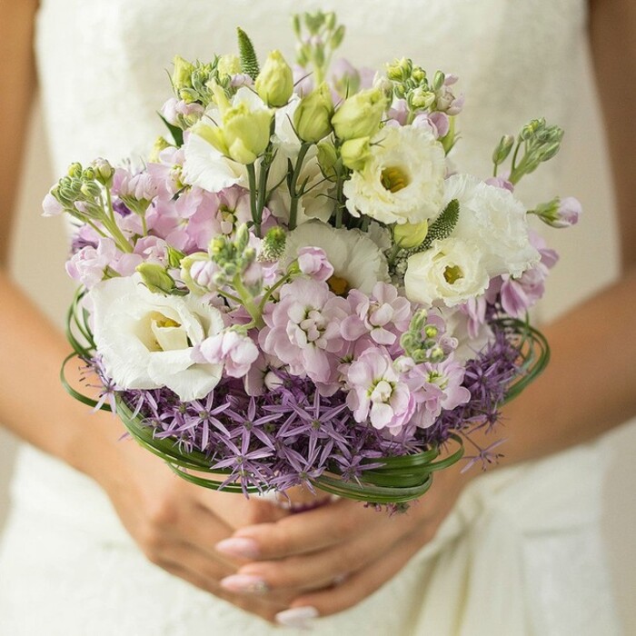 The artistry of Canonbury Flowers shines through a detailed eye-level close-up showcasing a bridal bouquet held gently by hands adorned with subtle pink nails-posed perhaps just outside a classic Islington townhouse. This bouquet is a masterclass in contemporary wedding floristry; white Lisianthus blossoms, open and expressive, take pride of place at the heart of the arrangement, their petals edged delicately and centers kissed with yellow-green. Encircling these are buds yet to bloom and richly textured clusters of pale pink and lavender stock, whose fragrance mingles softly in the morning air near the canal. A distinctive, spiky collar of purple Allium forms the bouquet's base like a modern art flourish, juxtaposed beautifully against the softness above. Bands of deep emerald bear grass spiral through the middle, their shine and structure recalling the lush greenways lining the edges of Canonbury's neighbourhood parks. The overall impression is one of gentle celebration and warmth, underscored by the bride's elegant white gown-its suggestion of lace visible in the softly blurred background. Crafted for a spring or summer wedding, this bouquet is not only a visual delight but an aromatic, tactile expression of love, freshness, and meticulous local craftsmanship-an ideal reflection of both classic elegance and contemporary Canonbury flair.