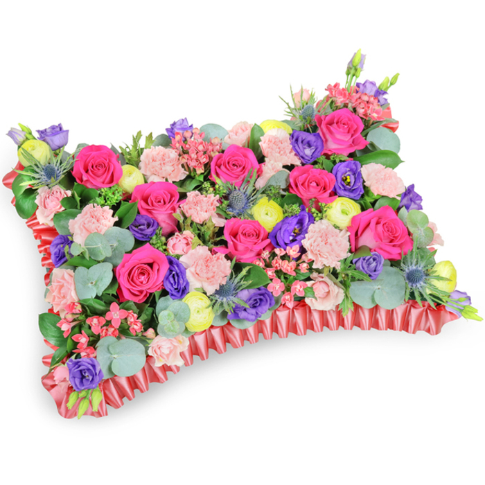 Photographed at a slight angle from above on a pure white background, this rectangular floral pillow reads like a hand-stitched tapestry of colour and texture, densely populated with fresh blooms and foliage. Hot-pink roses take centre stage - some tight and perfect, others loosening into velvet layers - surrounded by clusters of pale pink carnations whose frills create a soft, tactile surface. Intermittent deep-purple lisianthus blooms give pockets of cool depth, while lemon-yellow ranunculus act as sparkling accents across the arrangement. The inclusion of silver-blue eryngium thistles introduces an architectural, almost wild element that contrasts beautifully with the roses' softness. Eucalyptus leaves with their round, silvery-green faces and other leafy greens weave through the cushion, adding dimension and a faint, refreshing scent that calms the composition. The entire piece is set upon a rich, ruffled red satin border, pleated into a subtle frill that clearly defines the pillow's shape and lends a refined finish. Gentle, diffused light reveals the varied textures - from the smooth, satiny ribbon to the delicate carnation edges and the thistles' bristly silhouette - without distracting shadows. This arrangement is designed as a comforting sympathy or memorial cushion, offering warmth and celebration of life rather than sombre restraint; the lively mix of fuchsia, violet, lemon and blush reads as an embrace for grieving families. Crafted with local care by Canonbury Flowers, it is the kind of tribute often requested for funerals and memorial services in and around Canonbury Square and nearby Upper Street chapels.