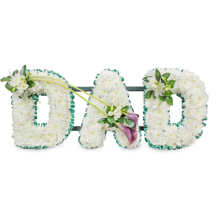 In this detailed funeral arrangement, the word DAD is formed in large, freestanding block letters, each one heavily padded with pristine white flowers that give a sense of softness and care. Every surface is densely filled with white chrysanthemums, their small, rounded petals overlapping to create a uniform, cushion-like texture that feels gentle to the touch even from a distance. The tribute sits against a pure white background, allowing the form and craftsmanship to stand out clearly, much as it would when displayed in a quiet chapel in Canonbury or during a memorial service in nearby Islington. A deep green satin ribbon, neatly ruffled and gathered, traces the outer edge of each letter, adding structure and a natural contrast, echoing the leafy calm of New River Walk. Faint glimpses of a dark support frame can be seen between the letters, indicating that the piece is constructed on a sturdy base designed to remain upright throughout the ceremony.   

Subtle accent arrangements bring extra character and emotion to the design. The upper left side of the first D is embellished with a dainty cluster of white freesia blossoms and fresh green foliage, their slender buds and open petals providing texture and a suggestion of fragrance. From this point, two long, arching green stems sweep diagonally across to the central A, where they anchor a more dramatic focal spray. Here, two deep purple calla lilies, with their smooth, sculpted trumpets, emerge from a bed of white freesia and polished leaves, providing a rich, solemn accent that speaks of dignity and deep affection. On the final D, a smaller spray of white flowers and greenery mirrors the first, completing the composition without overwhelming the simplicity of the word.   

The result is a moving, traditional tribute that many Canonbury families might choose when words feel inadequate: a floral DAD that quietly expresses love, respect, and remembrance at a funeral, graveside farewell, or celebration of life.