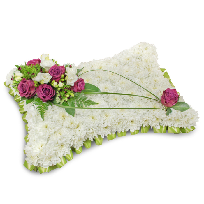 This carefully crafted pillow-shaped floral tribute is photographed on a plain white background so every texture reads clearly: a soft shadow anchors the piece and the simple setting allows the flowers' shapes and tones to speak. The surface is completely covered with compact white chrysanthemum florets, hundreds of tiny petals packed to form a plush, cloudlike cushion that evokes stillness and purity. A folded lime-green satin ribbon trims the entire perimeter in a tidy, scalloped ruffle that gives the cushion a finished, serene edge and a faint satin gleam. In the top-left quadrant a composed spray of deep fuchsia roses commands attention; their velvety petals open in luxuriant spirals and are gently offset by small white freesia or creamy lisianthus, introducing a delicate scent and lightness. The floral cluster is cushioned by abundant green foliage-feathery fern fronds and glossy leaves-that adds depth and natural contour. Across the white chrysanthemum field, two or three dark green bear-grass stems arc in elegant curves, their linear sweep lending motion and guiding the eye to the opposite corner where a single fuchsia rose mirrors the main cluster, quietly completing the arrangement. The overall impression is one of tender reverence: the textures of velvet, satin and soft chrysanthemum combine with subtle fragrance and soft studio lighting to create a tribute intended to comfort mourners. This pillow is a classic funeral choice for families in Canonbury and Islington, a composed expression of rest, solace, and remembrance suitable for chapel displays or graveside services.
