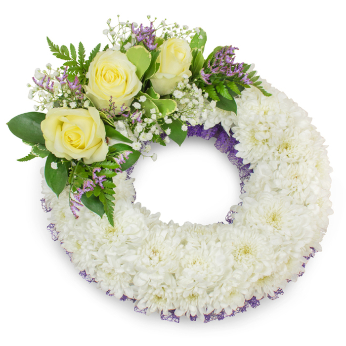 Presented on a stark white backdrop, this circular funeral wreath is a study in composed restraint and floristry skill. Its principal surface is formed from hundreds of small, snow-white chrysanthemums tightly packed to create a uniform, plush texture - each tiny bloom contributing to a cushiony, almost textile-like field that symbolizes peace and continuity. A slender, muted purple mesh or ribbon peeks intermittently from beneath the outer and inner rings of chrysanthemums, a tasteful accent that adds depth and a hint of sombre colour without disturbing the wreath's calm. Anchoring the piece in the upper-left quadrant is an arranged cluster of three creamy yellow roses, their multi-layered petals opening softly to express warmth and remembrance; these roses are surrounded by froths of white gypsophila and interlaced with small sprays of lavender-purple statice or limonium which introduce a refined contrast and delicate texture. The green elements are chosen for both contrast and motion: broad, waxy leaves provide a dark, steady backdrop while fine fern fronds and slimmer foliage infuse the composition with gentle, living movement. Soft, natural lighting reveals the cool tonality of the whites and the tender glow of the creams, casting a faint shadow that gives the wreath dimension and presence. As a sympathy arrangement crafted for families in Canonbury and the surrounding Islington neighbourhood - suitable for use at wakes, memorials, or church services near Canonbury Square, New North Road or St Paul's Church - it reads as a composed expression of condolence, offering a tactile, visual moment of solace and respectful remembrance.