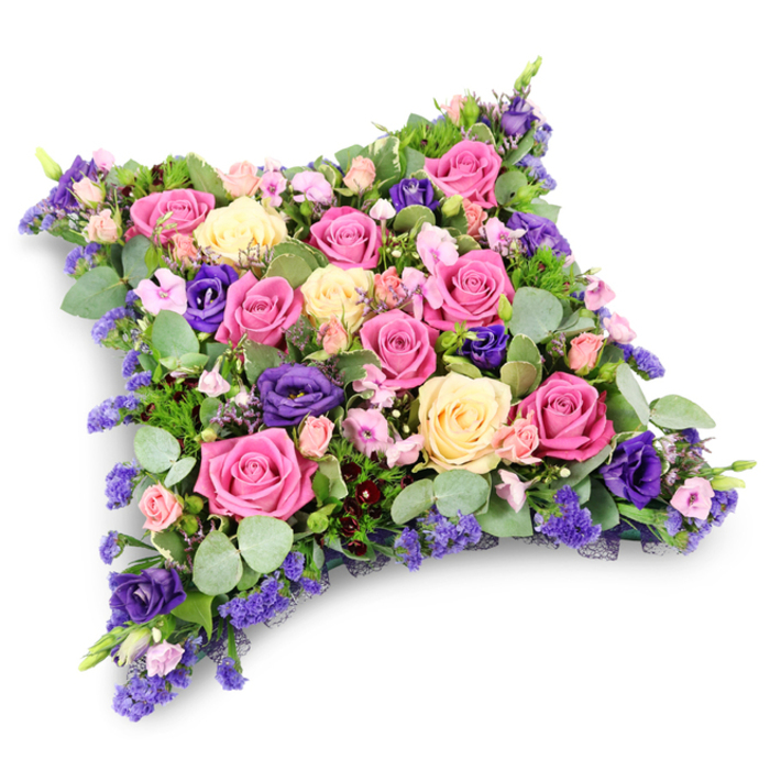 A beautifully crafted floral cushion, designed as a tribute for a funeral or memorial, appears almost to float against a crisp white background, its faint shadow emphasizing the soft, pillow-like shape. The arrangement forms a neat rectangle with rounded corners, fully carpeted in close-knit blooms that leave no gaps, only a continuous surface of colour and texture. Dominating the design are classic roses in varying tones: some a bright, confident fuchsia; others soft pastel pink; and a scattering of creamy pale yellow and ivory heads. Many roses are at their perfect stage of opening, with petals unfurling gently at the centre, creating intricate rosettes that catch the light. Between these, rich purple lisianthus blooms offer a velvety contrast, their deeper hue evoking quiet reflection and dignity. Interlaced among the main flowers are clusters of small lavender and lilac filler blossoms, reminiscent of statice or limonium, which bring a delicate, feathery finish to the surface and make the tribute feel even more plush. The edges and inner spaces are cushioned by a tapestry of greenery: rounded, silver-green eucalyptus leaves mingle with darker, fine-textured foliage that may be fern or similar greens, building a layered, natural frame around the roses. The overall impression is one of calm, respectful remembrance, akin to the gentle stillness of a Canonbury side street early in the morning. This floral pillow would be fitting for a service held in a local church or crematorium, offering a tangible symbol of comfort and eternal rest, crafted with the attentiveness you would expect from a long-established Canonbury florist. It embodies tenderness, care, and a quiet promise that a loved one's memory will be held close.