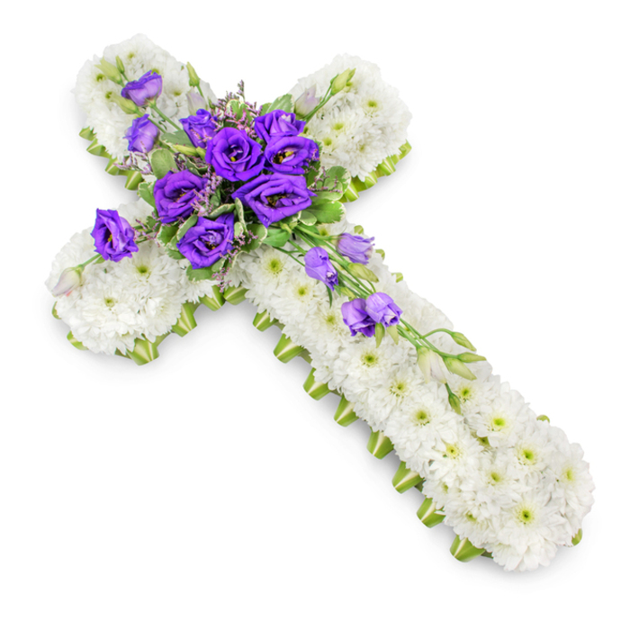 A traditional funeral cross, composed entirely of fresh blooms, is laid out on a pure white studio background where every nuance of the design is clearly visible. The cross itself is shaped from hundreds of white spray chrysanthemums placed so closely that they create a continuous, cushion-like surface. Their fine petals form small domes of softness, and the subtle green centres peep through, adding gentle depth without interrupting the tranquil white impression. Running along the outer sides of the cross is a precise band of bright lime-green ribbon, neatly folded around the floral base. This tidy edging frames the piece, echoing the fresh greens of nearby New River Walk and lending a contemporary accent to the otherwise classic tribute. At the junction where the arms of the cross meet, a bold arrangement of deep purple lisianthus commands attention. These flowers are rich and velvety, their layered petals spiralling inwards and catching the light to reveal hints of indigo in the shadows. Some blooms are wide open, others partially unfurling, and a few still in tight bud, creating a sense of natural progression and life. Interspersed among them are light sprigs of greenery and small filler flowers that break up the colour and add airiness. Several lisianthus stems extend from this central cluster, trailing diagonally down the lower-right arm, as if gently cascading in a soft, flowing line. A faint, diffused shadow sits beneath the cross, giving the tribute a slight lift from the surface. This floral cross, so suitable for a Canonbury church service or a quiet farewell at Islington crematorium, expresses purity, devotion, and heartfelt sympathy in a calm, dignified manner.