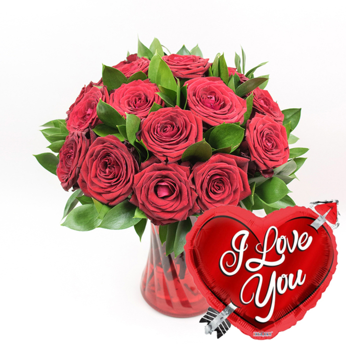 A richly romantic still-life unfolds against a crisp, white background: a lush bouquet of deep red roses, overflowing with texture and sentiment, arranged as though ready for same-day delivery to a loved one in Canonbury. Approximately a dozen large blooms gather at the heart of the design, their petals a luxurious velvet red that deepens towards the centre of each flower. Some roses are fully open, their petals curving backwards to reveal generous, rounded hearts, while others remain more compact, promising the slow, satisfying unfurling that stretches the gift over several days. Nestled throughout are sprigs of abundant dark green foliage, their surfaces smooth and slightly glossy, providing contrast and structure that feels as natural as the greenery in nearby Canonbury gardens and squares. The flowers sit in a stylish translucent red glass vase, the glass clear enough to hint at the stems within but richly tinted in ruby tones. Vertical ridges or subtle swirls run up the sides, catching the light and creating glimmers that echo the radiance of the blooms above. In the lower right foreground, overlapping the vase and drawing the eye immediately, is a shiny metallic red heart-shaped balloon. Its surface reflects soft highlights, and across its centre the words I Love You appear in bold, flowing white script, instantly legible. A silvery arrow, reminiscent of Cupid's, seems to pierce the heart from lower left to upper right, adding a playful, iconic touch. The restrained white background allows the reds and greens to command attention, giving the whole piece the feeling of a carefully considered romantic gift-perfect for anniversaries, proposals, or Valentine's surprises in and around Canonbury.