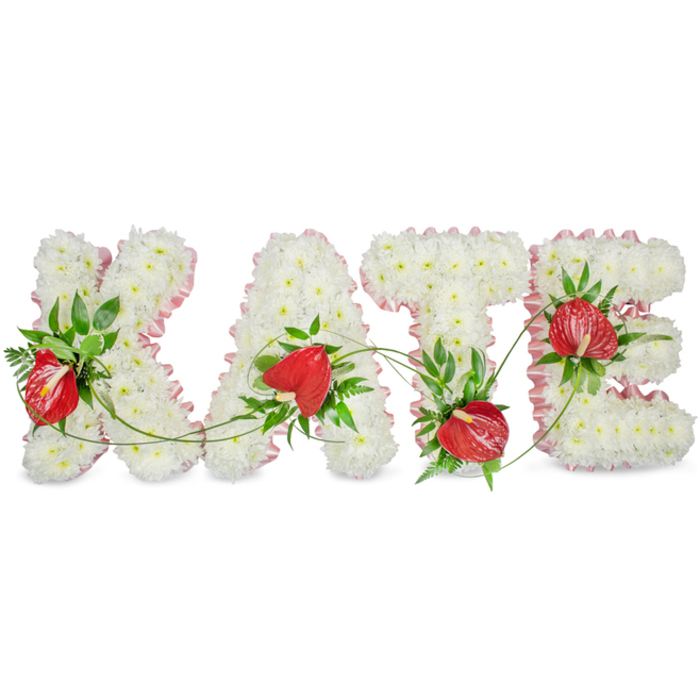 A large, three-dimensional memorial display spells KATE in capital letters, each letter formed from a continuous, uniform carpet of small white chrysanthemums that create a soft, textured surface reminiscent of freshly fallen snow. The outer edges of the letters are defined by a delicate pale pink ribbon or fabric trim, its slight ruffle adding a soft outline and a gentle warmth against the immaculate white blooms. Vibrant red anthuriums are artfully positioned within the white bases, their glossy, heart-shaped spathes and central spadices offering luminous focal points that draw the eye across each letter. Interwoven around the blooms is an abundance of dark green foliage - broad glossy leaves, fern fronds, and sprigs that lend depth, contrast, and a verdant scent. Fine green trailing vines snake gracefully between the A and the T and bridge the negative space, reinforcing the sense of connection across the name. Set against a bright white background with a faint drop shadow, the letters appear to float slightly above the surface, a subtle three-dimensional effect that adds dignity and presence. This piece reads as a considered funeral tribute, the pairing of classic white chrysanthemums and bold red anthuriums conveying both purity and heartfelt warmth. A florist in Canonbury or on Essex Road might prepare such a bespoke tribute for a memorial service at a local church or a celebration of life at a nearby community venue, presenting an arrangement that balances technical craft, seasonal freshness, and the quiet poetry of remembrance.