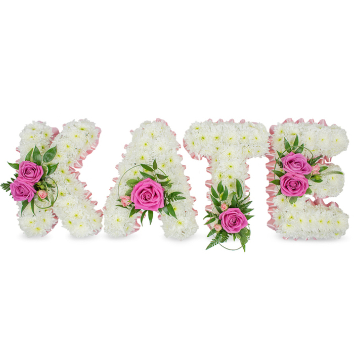 This bespoke floral composition spells the name KATE in substantial, freestanding block letters, each letter meticulously constructed from countless tiny, pure-white chrysanthemums to form a dense, pillowy canvas. A soft, pale-pink satin ribbon traces the inner edges of every letter, lending a faint blush that gently separates the white expanse and adds a feminine, heirloom quality to the design. Hot-pink roses function as emotional anchors: two gracefully opened roses sit on the left arm of the K, a single, commanding pink rose nestles in the low central bowl of the A, a solitary vivid rose punctuates the base of the T, and two roses cluster along the right side of the E. Intertwined are rich green elements - fern-like fronds, ruscus and variegated leaves - together with tiny pale-pink filler blooms that create soft transitions and a subtle, garden-fresh scent. Set against an immaculate white backdrop the letters cast soft, grounding shadows, giving the tribute a sculptural presence as if it might be placed at a chapel doorway or beside a commemorative photograph. The dense packing of chrysanthemums yields a cloud-like texture that invites touch, while the roses provide a satin-petalled contrast and a gentle perfume. This arrangement reads as a deeply personal expression of love, sympathy and remembrance, appropriate for memorial services or a celebration of life in the Canonbury area - quietly resonant for families meeting near Canonbury Square, Essex Road or along New River Walk.