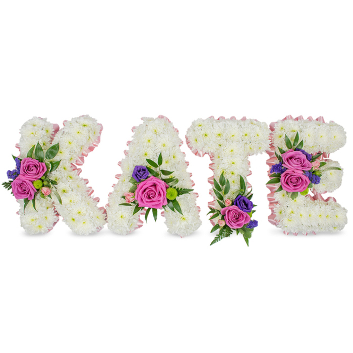 A poignant, personalised floral name tribute reading KATE, composed as four freestanding letters formed entirely from compact, snow-white chrysanthemums that produce a dense, silky texture and a sense of serene purity. Each letter is edged with a subtle pale pink satin ribbon that gently defines the shapes and lends a soft, blushing outline. Carefully positioned accent bouquets enliven the white base with saturated colour: large, velvety magenta-fuchsia roses sit with smaller pastel pink spray roses, and fine stems of deep purple or blue accent flowers - likely statice or lisianthus - for contrast. The foliage choices, from airy fern fronds to smooth eucalyptus leaves, provide layered green textures and a faint, cooling scent. Placement of the accent clusters follows a considered pattern - the K bears a bold cluster on its diagonal arm; the A displays blooms near its bottom right curve; the T is grounded by a grouping near its base; and the E receives two balanced clusters on the middle bar and lower arm - giving the whole piece a harmonious rhythm when viewed from the front. Under soft, neutral lighting on a white background, the bright floral pops read as comforting rather than celebratory, making this arrangement especially suited to memorial services, graveside tributes or a chapel setting in Islington, near Upper Street or Canonbury's quieter lanes. The workmanship shows the floristry tradecraft of a neighbourhood maker: considered, restrained and deeply respectful, designed to convey sympathy and remembrance.