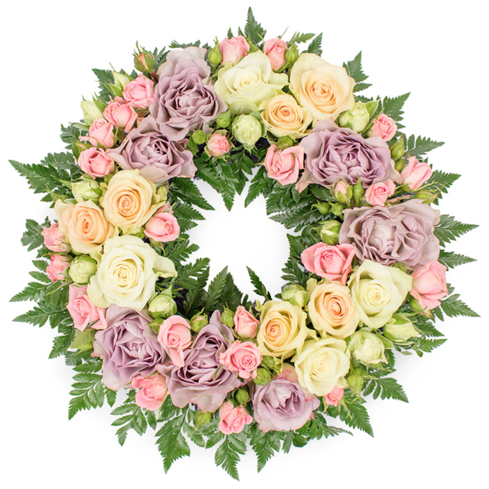The image presents a generous, circular funeral wreath placed against a pure white backdrop, allowing every nuance of its pastel roses and dark green foliage to stand out with quiet clarity. The wreath is densely composed, so that its structure is entirely concealed beneath a thick carpet of blooms and leaves. Large, fully opened roses in soft ivory and creamy white form the foundation, their petals layered like silk, gently curling back to reveal velvety centers. Interlaced with these are smaller clusters of blush pink spray roses and tightly furled buds, the varying stages of bloom echoing the passage of time and the different chapters of a life remembered. Subtle notes of pale peach and buttery yellow roses infuse the design with a gentle warmth, while distinctive dusty lavender and muted mauve roses introduce a refined, antique elegance often chosen for thoughtful sympathy tributes. The blossoms rest on a verdant base of dark green fern fronds, their fine, feathery leaves radiating outward and occasionally peeking through the flowers, adding depth, structure, and a cool forest freshness. The wreath's surface rises in a slight dome, catching light across the petals and casting soft shadows that enhance its dimensionality. It feels like the sort of carefully considered design a Canonbury family might select from a trusted local studio for a service near New River Walk, or for a memorial gathering in a townhouse just off St Paul's Road. Radiating calm, respect, and enduring affection, this handcrafted wreath embodies a tender farewell and offers visual comfort to those gathered in grief.