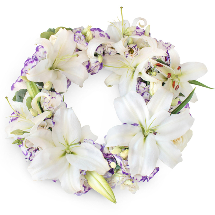 A meticulously arranged circular sympathy wreath rests against a pure white background, its refined mix of white and purple flowers capturing a quiet, reverent beauty. The focal blooms are generous white Oriental lilies, their petals gently recurved and almost luminous, creating soft shadows that suggest depth and softness. Many lilies have fully opened to show their pale green centres and fine, amber-tipped stamens, while a few remain in half-opened stages, their promise of bloom echoing the ongoing cycle of life and memory. Interwoven throughout the wreath are elegant Lisianthus-like blossoms whose white petals are finely brushed or fringed with lilac and gentle violet, the ruffled edges adding a cloud-like texture between the more architectural lilies. Nestled here and there are small buds and possibly tiny, cream-toned rosebuds, each one like a whispered farewell or private thought. Beneath the flowers, rich green foliage and subtle grey-green sprigs reminiscent of eucalyptus ripple softly around the circle, suggesting the kind of greenery one might see in a secluded Canonbury courtyard or along the quiet lanes near New River Walk. The ring is full yet not overbearing, creating an unbroken halo of flowers that symbolises eternal love, purity, and the continuity of the soul's journey. Even, diffused light ensures the whites remain crisp and the purples gentle, with no harsh glare, reflecting the calm environment of a thoughtful farewell. This wreath would be a fitting choice for a funeral or memorial held in and around Canonbury, a comforting floral tribute placed beside the casket or memorial table to honour a cherished life with grace and tenderness.