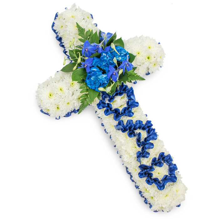 This image shows a substantial funeral flower cross laid on a clean, bright white background, photographed so that every petal and ribbon fold is clearly visible. The entire cross is densely packed with small white spray chrysanthemum heads, placed closely side by side until no base is visible. Their petals form a soft, textured surface that feels almost like a quilt of flowers, creating a strong sense of peace and purity. Tracing the outer contour of the cross is a band of royal blue satin ribbon, gently ruffled, with a subtle sheen that catches the light along its edges. This deep blue border defines the shape crisply and adds a formal, almost ceremonial tone that suits a funeral or memorial service in the Canonbury and Islington area. At the heart of the cross, where the arms meet, sits a prominent collection of vibrant blue blooms: frilled blue carnations or dyed chrysanthemums, paired with slimmer, upright stems that resemble twisted spikes of blue delphiniums or orchids. These flowers vary in tone from dark ink-blue to lighter azure, giving depth when seen against the white background. Around them, glossy green foliage and delicate fern tips introduce movement and a touch of natural woodland calm. Along the centre of the vertical stem, the word DAD is artistically formed using loops and folds of the same royal blue ribbon, each letter standing out clearly from the white chrysanthemums below. The lighting is even and shadow is minimal, making this bespoke floral cross a clearly legible, deeply personal tribute that would feel perfectly at home in a quiet Canonbury church, chapel of rest, or graveside committal for a cherished father.