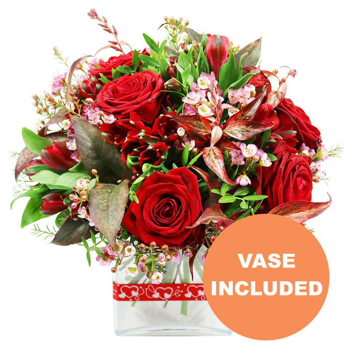 An opulent bouquet of rich red blooms is presented against a pristine white background, its rounded dome of flowers reminiscent of a carefully composed centrepiece ready to brighten a Canonbury townhouse near New River Walk. At its core, sumptuous red roses with velvety, tightly layered petals cluster together, some fully open in luxurious spirals, others half-unfurled in a state of poised anticipation. Surrounding these star roses, smaller deep crimson flowers similar to alstroemeria cluster in sprays, their multiple petals introducing a fine, almost feathery texture that contrasts with the bold rose heads. Between the red petals, dainty stems of pale pink and white waxflower or limonium scatter tiny blossoms like soft, scented stars, adding luminosity and airiness to the design. The bouquet is cradled by a generous wreath of foliage: broad, glossy green leaves lend structure and freshness, while lighter, more delicate sprigs soften the profile and create a sense of movement. Here and there, dramatic burgundy and reddish-brown leaves with subtle variegation arc outward, deepening the colour story and giving the arrangement a sophisticated, evening-appropriate elegance that feels at home in Canonbury's period drawing rooms and contemporary apartments alike. The stems are gathered into a clear glass rectangular vase with smooth, modern lines, its transparency allowing the viewer to see the cut stems and clean water inside, reinforcing the sense of freshness. Around the lower portion of the vase, a wide red ribbon printed with crisp white heart outlines wraps neatly, its playful, romantic motif suggesting Valentine's surprises, anniversary dinners, or a heartfelt apology. In the lower right corner of the image, a bold circular orange emblem contains bright white uppercase lettering reading VASE INCLUDED, making it clear that this striking, ready-to-gift bouquet arrives complete, ideal for convenient flower delivery throughout Canonbury and the wider N1 area.