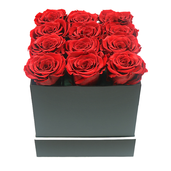 This image presents a meticulously styled floral gift that feels refined and intentional, the kind of design a Canonbury florist might recommend for a landmark anniversary or a quietly grand romantic gesture. At the centre is a square gift box in deep, matte black, its surfaces smooth and unadorned except for a thin, sharp white band that wraps around the lower part like a delicate belt, accentuating its tailored, boxy silhouette. Resting within the box are twelve exquisite preserved roses, arranged in three neat rows of four so that their circular heads form a precise grid. Each rose is a saturated ruby red, with petals that appear thick and velvety, curling outwards and then folding back on themselves in intricate spirals. The blooms are fully open and uniform in height, forming an even, softly undulating surface that lifts slightly above the rim, as if the roses are gently overflowing without actually spilling beyond the edges. The lighting is soft and controlled, creating a gentle sheen on the outer petals and faint shadows between the blooms that emphasize their depth and texture. There are no leaves, stems, or additional decorations, allowing the simple colour palette of black, white, and red to make a strong, modern statement. The background is pure white, giving the image a clean, editorial quality while drawing attention to every curve and contour of the flowers and the geometric precision of the box. The arrangement suggests enduring romance and thoughtful sophistication-something you might send to a loved one living along Canonbury Road, or display as a lasting centrepiece in a calm, minimalist living room overlooking New River Walk.