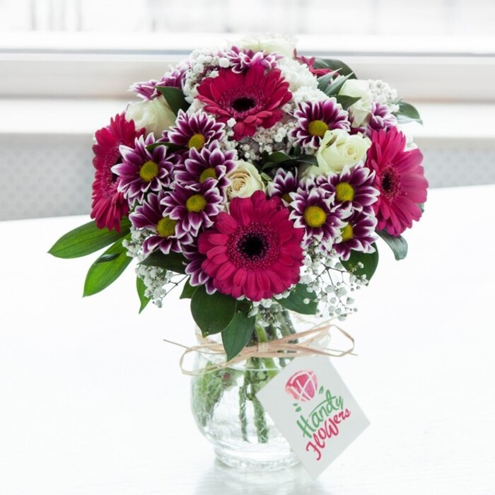 A lush, professionally arranged bouquet of mixed flowers is showcased in the centre of the frame, resting in a clear, textured glass jar-style vase against a bright, softly diffused white background, like a sun-washed ledge in a Canonbury townhouse. Dominating the design are generous gerbera daisies in vivid fuchsia and magenta tones, their broad petals radiating from deep, inky centres that lend dramatic contrast. Nestled around them are intricate bicolour chrysanthemums, each bloom displaying petals that graduate from rich purple at the edges to clean white nearer the centre, surrounding a bright yellow eye that seems to shimmer in the natural light. Among these bold tones, creamy white spray roses appear in different stages of opening, from tight buds with smooth, waxy petals to softly unfurled blossoms, bringing a refined, romantic calm to the overall palette. Fine sprays of white gypsophila thread delicately through the bouquet, their tiny blossoms catching the light and creating a gentle, cloud-like veil between the larger flowers. Deep green foliage supports and frames the arrangement, leaves overlapping and arching outward in a way that adds structure while keeping the look airy and fresh. The stems are visible through the vase's clear glass, neatly cut and fanned in a spiral, held in place by clear water that hints at just-cut freshness, as though these flowers were arranged moments ago in a local Canonbury studio. Around the neck of the vase, a length of rustic twine is tied into a simple bow, from which hangs a small, white rectangular gift tag bearing a stylised pink blossom and delicate writing, ready to carry a personal message. The overall impression is of a joyful yet elegant bouquet, perfect for same-day delivery to homes near Canonbury Square or along New North Road, bringing an immediate sense of celebration and warmth to any room.