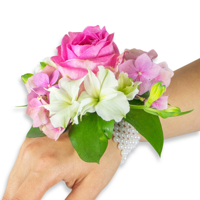 Seen against a stark white studio background, this elegant wrist corsage rests on the left forearm of a person with a light-to-medium skin tone, every detail sharply lit to reveal texture and form. The composition is centred on a large, fully opened hot fuchsia-pink rose whose dense, overlapping petals create a sumptuous, tactile focal point. Surrounding the rose, clusters of blush-pink hydrangea florets offer soft, rounded volume and a whisper of papery crispness, while several white alstroemeria - Peruvian lilies - are artfully interspersed, their smooth petals edged subtly with pale green veining that draws attention to each flower's centre. On the outer right of the arrangement, a small grouping of light green alstroemeria buds and a scattering of barely opened pink buds hint at the promise of further bloom, adding depth and a sense of time. Rich emerald leaves provide a deep, cooling frame, their broad surfaces grounding the pastel palette. The corsage is secured to the wrist by an elegant multi-strand bracelet crafted from tiny, lustrous white pearls; the strands form a delicate net that holds the bouquet in place and extends partly onto the back of the hand, combining floral artistry with jewellery-like restraint. The overall mood is classic and romantic - perfect for a wedding, prom, or formal reception - and suggests the careful hand of a local florist who knows the quiet charm of Canonbury, from Essex Road markets to the shaded walks by New River.