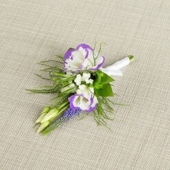 A carefully composed floral boutonniere lies against a backdrop of light beige linen, the subtle weave of the fabric giving a soft, tactile context to the elegant flowers. Dominating the arrangement are two open lisianthus blooms, their white petals gently curled and richly ruffled at the edges in a vivid purple that echoes the twilight tones often seen over Canonbury's rooftops. Nestled against them is a slender stalk of muscari, its tiny, bead-like blossoms forming a compact column of deep violet-blue that adds both depth and a touch of whimsy. Threaded between these focal flowers are tiny white filler blooms, reminiscent of baby's breath, which create a halo of softness and suggest the fine mist of petals you might see in a florist's studio along Newington Green. Wisps of feathery green foliage spread out around the cluster, light and airy, their fine texture evoking the gentle brush of grasses in nearby parks. Two pale green buds, still tightly closed, sit within the composition, hinting at flowers not yet unfurled and the promise of moments still to come. The stems are neatly aligned and wrapped in a smooth band of white satin ribbon, finished with a modest bow that signals this is a piece crafted for a significant occasion-a wedding, civil ceremony, or formal event. The light feels natural and calm, highlighting the fresh, dewy quality of the petals as if they were arranged earlier that morning by a Canonbury florist preparing for same-day delivery to a ceremony venue tucked away off Essex Road. The overall impression is one of quiet anticipation, elegance, and thoughtful design.