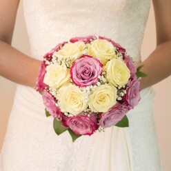 In the heart of Canonbury, this close-up of a bridal bouquet tells its own love story-one delicately composed of creamy lemon-yellow roses nestled against lively fuchsia blooms, each hue enhanced by the expert touch of a seasoned florist. Subtle notes of mauve and lavender pink grace some petals, adding a gentle complexity reminiscent of Canonbury's diverse gardens. Wisps of fine white gypsophila are interlaced throughout, lending the bouquet an airy lightness that seems to float above the bride's hands, which cradle the arrangement with quiet reverence. Dark green leaves peek from beneath, echoing the abundance found along local pathways like Canonbury Grove. The bride's ivory gown, adorned with lace featuring elegant floral and swirling motifs, catches the soft natural light, echoing the detailed beauty of the bouquet. Each flower is showcased by the even brightness, freshness radiating from every petal and lace fold. The composition captures a moment of celebration, steeped in the quiet elegance unique to Canonbury, evoking images of intimate ceremonies in neighborhood churches or garden receptions by New River Walk. For anyone seeking floral inspiration for special occasions, this bouquet mirrors a blend of timeless romance, artistry, and the vivid local character found only in Canonbury.