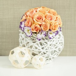 On a smooth, white tabletop that could just as easily belong in a Canonbury dining room as in a wedding venue, a graceful floral centerpiece becomes the quiet focus of the scene. The arrangement is built around a neatly sculpted globe of roses in shades of soft peach and warm apricot, each flower at its peak, petals unfurling in generous layers that give a plush, cushioned effect. There is a sense of calm abundance, as if every rose has been placed with care to create a perfectly even, velvety dome. Encircling the base of this rose cloud is a ring of white lisianthus blooms, their frilled petals finely edged with vivid purple, adding contrast and a touch of modern flair. Sprigs of tiny white gypsophila peek between them like stars against a twilight sky, softening transitions and lending a floating lightness. The flowers rise from a low, broadly spherical white basket-vase, woven from firm rattan or similar natural strands. Its open, crisscross pattern and bulbous silhouette echo the roundness of the blooms above, while the bright white finish keeps the look crisp and contemporary, ideal for a Canonbury Flowers wedding table along nearby Upper Street or a stylish dinner party at home. In the foreground, two pale wooden decorative balls, one medium and one smaller, rest on the white surface, their airy latticework echoing the container's craftsmanship. Behind it all, a gently textured beige fabric backdrop blurs away, allowing the soft peaches, violets, and whites to stand out. The mood is serene, romantic, and thoughtfully composed, perfect for celebrating milestones or styling a refined reception table.