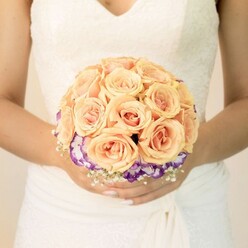 This image focuses tightly on a bride from the waist up, standing against a softly blurred, pale background that might be the quiet interior of a Canonbury townhouse or the light-filled room of a local registry office. Her wedding gown is a classic ivory, with lace tracing the bodice and a subtle V-shaped neckline, the skirt flowing in soft folds that frame, but never distract from, the bouquet she holds. Cradled in both hands, the round bridal bouquet is a carefully composed dome of roses in gentle shades of peach, apricot, and blush pink, each bloom densely petalled and almost porcelain in appearance. The roses are clustered closely together to create a smooth, cushion-like surface, their colours blending like a warm sunrise. Below this main cluster, a striking ring of deep purple blooms with ruffled white edges-likely lisianthus-forms a contrasting collar that encircles the roses, giving the bouquet depth and elegant definition. Tiny white filler flowers, similar to baby's breath, are scattered delicately around the base, adding a fine, airy texture that lightens the overall silhouette. The bride's nails are neatly manicured in a light, neutral polish, with one thumb showing a hint of sparkle that catches the light, echoing the understated sophistication of the arrangement. The scene feels calm, intimate, and romantic, evoking the quiet excitement of a Canonbury wedding morning as flowers freshly delivered from a nearby florist rest cool against the bride's hands, ready to be carried down the aisle along tree-lined streets and through historic local venues.