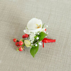 An artfully composed boutonniere lies centred on a pale beige, woven linen surface, its neutral background allowing every floral detail to stand out clearly, as if set down on a carefully pressed cloth in a quiet Canonbury mews house moments before a ceremony. The focal point is a flawless white Phalaenopsis orchid, its wide, satin-smooth petals curling gently outwards to reveal a soft, buttery-yellow and muted orange throat that adds depth and gentle warmth. Surrounding this central orchid is a light cloud of baby's breath, each tiny white floret forming a lace-like halo that softens the design and introduces a sense of airiness and romance. Carefully placed around the flowers, sleek green leaves in different lengths and shapes add structure and a fresh, woodland feel, echoing the greenery seen along the paths of nearby New River Walk. Tucked to one side is a vivid cluster of glossy, cherry-red berries, accompanied by a few pale green buds that suggest natural variety and seasonal interest, bringing a cheery, almost festive note to the otherwise restrained palette. The stems at the base are firmly bound with a rich red satin ribbon, wrapped tight and finished in a small, neat knot, its short tails lying flat and catching just a hint of light. This refined floral accessory, with its serene white, lush green, and celebratory red accents, feels perfectly suited for weddings, winter formals, and special gatherings across Canonbury and the surrounding Islington neighbourhoods, capturing a moment of calm anticipation before the day's events unfold.
