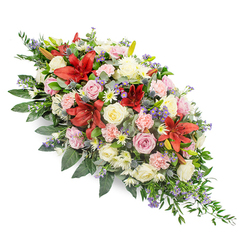 Set against a bright white backdrop, this elongated funeral spray appears almost to float, its colours and textures forming a gentle river of flowers from one end to the other. The composition is dense yet carefully balanced, beginning with a cluster of deep red lilies whose broad, star-shaped petals open confidently, their rich colour echoing the emotional depth of farewell. Around these, white and soft pink roses are arranged at different stages of bloom, from shy buds suggesting unspoken words to fully opened roses representing a life shared and remembered. Light pink carnations with their softly scalloped petals weave between the roses, joined by tiny white chrysanthemum or daisy-like florets that lend the whole tribute a sense of light and clarity. At intervals, cool blue-purple thistles emerge, their spiky forms and silvery tones adding a distinctive texture, while fine sprays of lavender-blue filler flowers contribute subtle hints of colour that never overwhelm. A generous collar of greenery grounds the design: broad, dark green leaves create depth and structure, while feathery, fern-like sprigs soften the edges and stretch outward, tapering with the overall form to a delicate point. The spray feels composed with quiet intention, the kind of thoughtful sympathy piece chosen for a funeral service in Canonbury, perhaps carried through the calm streets around Canonbury Square or set beside a memorial in a nearby church. It communicates love, respect, and gentle support, crafted in the style of a local florist who understands the need for beauty and composure on a difficult day.