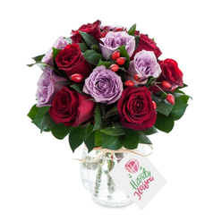 A richly hued, romantic bouquet of crimson and lavender roses, carefully formed into a rounded, abundant silhouette and set against a stark white field. Deep, velvety red roses - a mix of crimson and burgundy tones - sit shoulder to shoulder with soft mauve and lilac roses, their petals unfurling like pages of a well-loved book. The colours converse across the arrangement: the reds offer warmth and intensity while the lavender flowers introduce a calming, elegant counterpoint. Bright red-orange rose hips are dotted through the mass of blooms, their small, firm shapes adding a lively, almost playful punctuation and an organic, countryside texture that suggests hedgerows after rain. Rich, dark green ruscus or salal leaves weave through the bouquet, providing cool contrast and a subtle, leafy scent when the stems are handled. The flowers are presented in a rounded mercury glass vase with an antique silvered finish that catches light in soft, mottled reflections; natural raffia is tied at the neck to lend handmade charm. A white rectangular tag hangs from the twine, bearing a modest stylised rose emblem and space for a short note. Under soft, diffused lighting the petals appear plush and deeply saturated, the vase gleams gently, and the whole composition reads as thoughtful and refined. This floral piece evokes romance and quiet luxury - perfect for marking an anniversary, offering comfort, or sent as a deliberate, elegant gesture to someone special in Canonbury or nearby Islington neighbourhoods.