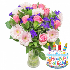 Bathed in soft, neutral light against a crisp white backdrop, this birthday arrangement blends classic romance with a touch of party-ready fun. The dominant element is a lush cluster of soft pink roses: petals layered like silk, ranging from pale blush at the edges to richer pinks toward the center, arranged to create a rounded, generous silhouette. Complementing the roses are pale pink Gerbera daisies whose broad petals and pale yellow centers open like friendly faces, giving the bouquet cheer and presence. Scattered throughout are concentrated sprays of vivid purple statice (limonium), their textured, papery blooms creating pockets of contrast and a painterly sense of depth. Small white filler blooms and abundant glossy green foliage support the composition, framing each flower and lending a natural, garden-fresh feel. The stems rest in a transparent glass vase with a slightly wider base so water and green stalks are visible, emphasizing freshness and careful handling. Nestled to the right, a cake-shaped foil balloon adds celebration: white frosting with wavy blue piping, colourful sprinkles, and four multicoloured candles-depicted purple, red, orange and blue with yellow flames-above a playful Happy Birthday message spelled in bright letters; a tiny KALEIDOSCOPE mark is visible at the bottom. As a Canonbury florist, I imagine sending this as a birthday surprise for neighbours near Canonbury Square or deliveries across Essex Road and Newington Green-an arrangement that reads as intimate, jubilant, and freshly prepared by Canonbury Flowers for a loved one's special day.