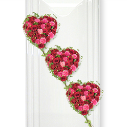 A trio of rose-filled hearts descends in a soft diagonal line, creating a powerful and tender funeral tribute that seems to gently drift down a pristine white wall. Each heart is formed from an abundance of roses, so closely set that no gaps show, only a luxurious carpet of petals in shades of deep crimson, vivid cerise, and delicate blush pink. The red roses at the centre of each heart feel rich and profound, their velvety petals opening in layered spirals that suggest both strength and vulnerability. Interspersed among them, the pink roses lighten the mood, echoing memories of warmth, kindness, and shared joy. Fine ivy vines edge the contours, their dark green leaves like small, glossy arrows pointing around each heart and then trailing down to touch the next, linking the three shapes into one continuous gesture of love. The smooth white background and subtle vertical lines resemble a calm gallery or a quiet Canonbury chapel interior, allowing the colours to appear almost luminous, as though the hearts are gently illuminated by soft overhead lighting. This arrangement feels especially suited to a memorial service or celebration of life in and around Canonbury, perhaps for a farewell at a local crematorium or a funeral in nearby Highbury or Barnsbury, where family and friends gather. The cascading hearts suggest generations connected across time, making this design a poignant choice for honouring a cherished partner, parent, or grandparent with roses that speak clearly of loyalty, romance, and undimmed affection.