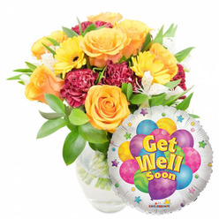 Soft daylight pours across a cheerful Get Well Soon arrangement that feels both handcrafted and distinctly local to Canonbury. A clear glass vase, water rippling gently around visible stems, supports a generous bouquet of open orange and golden-yellow roses; their petals are plush and slightly citrus-scented, forming a warm central focus. Nestled among them are sunny yellow gerbera daisies whose bold discs bring an optimistic, sunflower-like brightness, while rich burgundy carnations add plush texture and shadowed depth. Flecks of white alstroemeria weave through the larger blooms, lending delicate, papery contrast and a light, floral sweetness that keeps the palette airy. Abundant green foliage wraps the composition, leaves of varied shapes creating movement and freshness-imagine this piece arriving fresh from a morning run to Canonbury Square or a stop at the Essex Road market. Slightly overlapping the bouquet on the lower right is a shiny, round silver Mylar balloon that reflects the studio light; its playful centre shows a cluster of colourful cartoon balloons in blue, purple, pink, green and yellow, patterned with dots and stripes, and the phrase Get Well Soon displayed in cheerful yellow-and-blue letters. Small starburst accents mark the balloon's white border and a discreet KALEIDOSCOPE logo appears at the bottom edge. As prepared by Canonbury Flowers and intended for a bedside or hallway, this arrangement radiates encouraging warmth, fresh fragrance and thoughtful composition-perfect for sending comfort and quick-healing wishes across the local streets of Canonbury and nearby New River Walk.