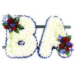 Carefully hand-crafted by Canonbury Flowers for families in and around Canonbury, this freestanding tribute spells the initials "BA" in bold, uppercase letters formed from hundreds of small, immaculate white chrysanthemums. The chrysanthemum heads are densely packed to create a soft, tactile surface that looks almost like carved cloud; their gentle texture invites a quiet touch while conveying a sense of purity and calm. Each letter is edged with a ruffled royal-blue satin ribbon that crisply defines the contours and offers a refined, ceremonial frame to the white base. The 'B' carries a lush accent on its upper left curve: several deep, velvety red roses in varying stages of bloom, paired with tall, cobalt-blue delphinium spikes that lend vertical contrast and a hint of coolness to the palette. Pale green tendrils and deeper green foliage weave through these flowers, adding movement and an organic finish. The 'A' mirrors this treatment with an equivalent cluster at its lower right, creating visual harmony across the pair. The two initials rest on a narrow, dark green horizontal stand that keeps them together as a single memorial statement. Photographed against a clean white backdrop and touched by soft, natural light - the sort of light you might find in a chapel on New River Walk or a small Canonbury chapel - the arrangement reads as dignified and intimate, a personalised floral message designed to honour memory and provide comfort.
